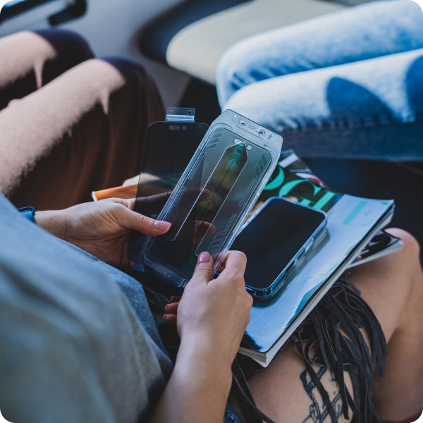 Szkło z funkcją prywatyzacji z aplikatorem trzymane w rękach. Na kolanach osoby trzymającej widoczny telefon